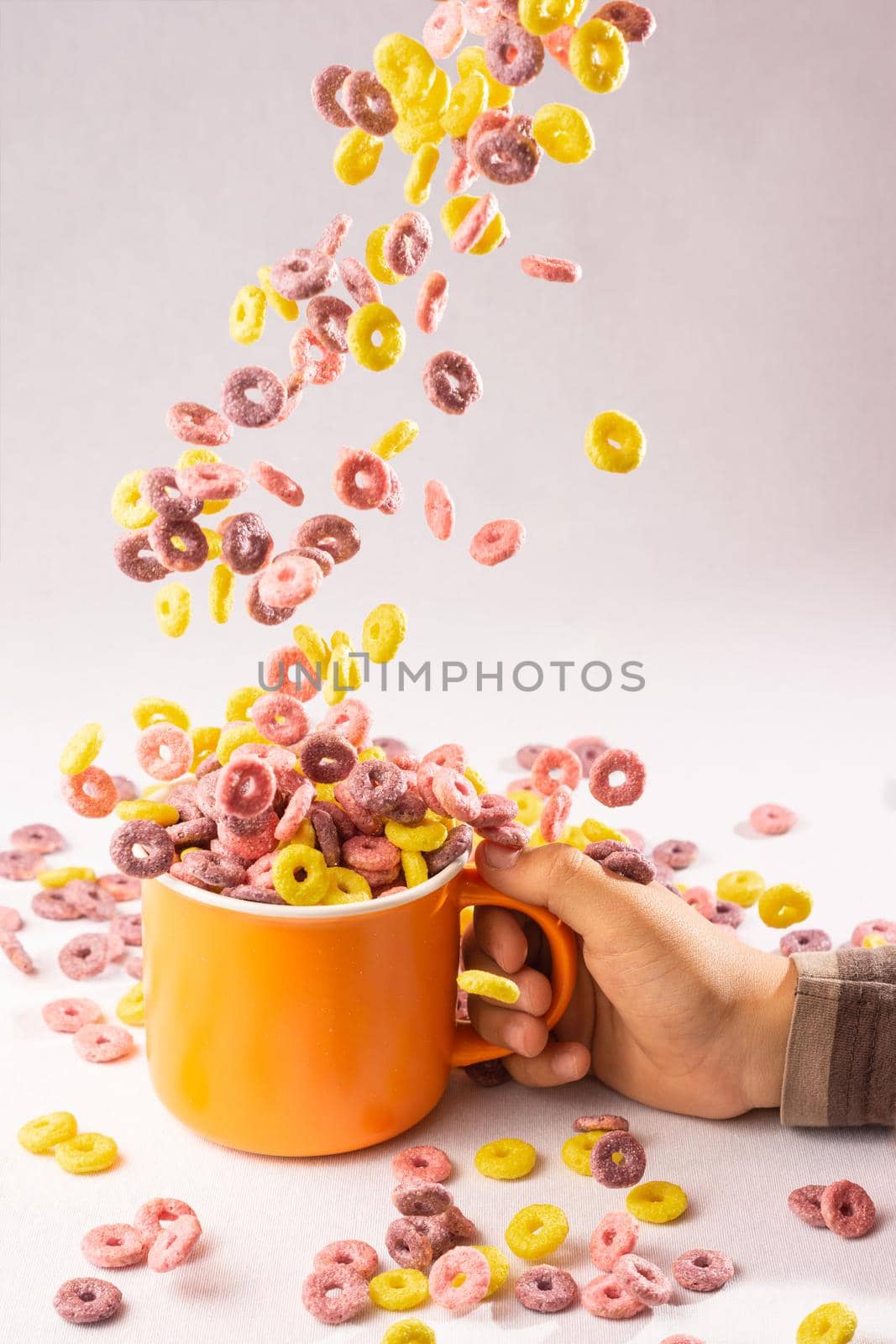 colored cereal rings falling into an orange cup held by a child's hand on white background with side lighting