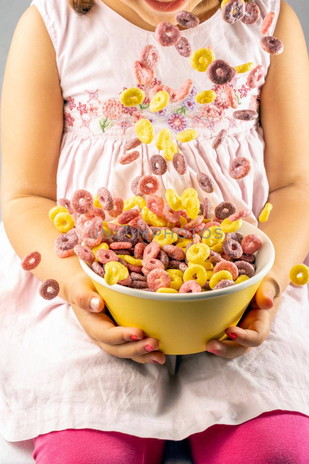 colored cereal rings falling into a yellow bowl held by a girl with both hands wearing a pink dress