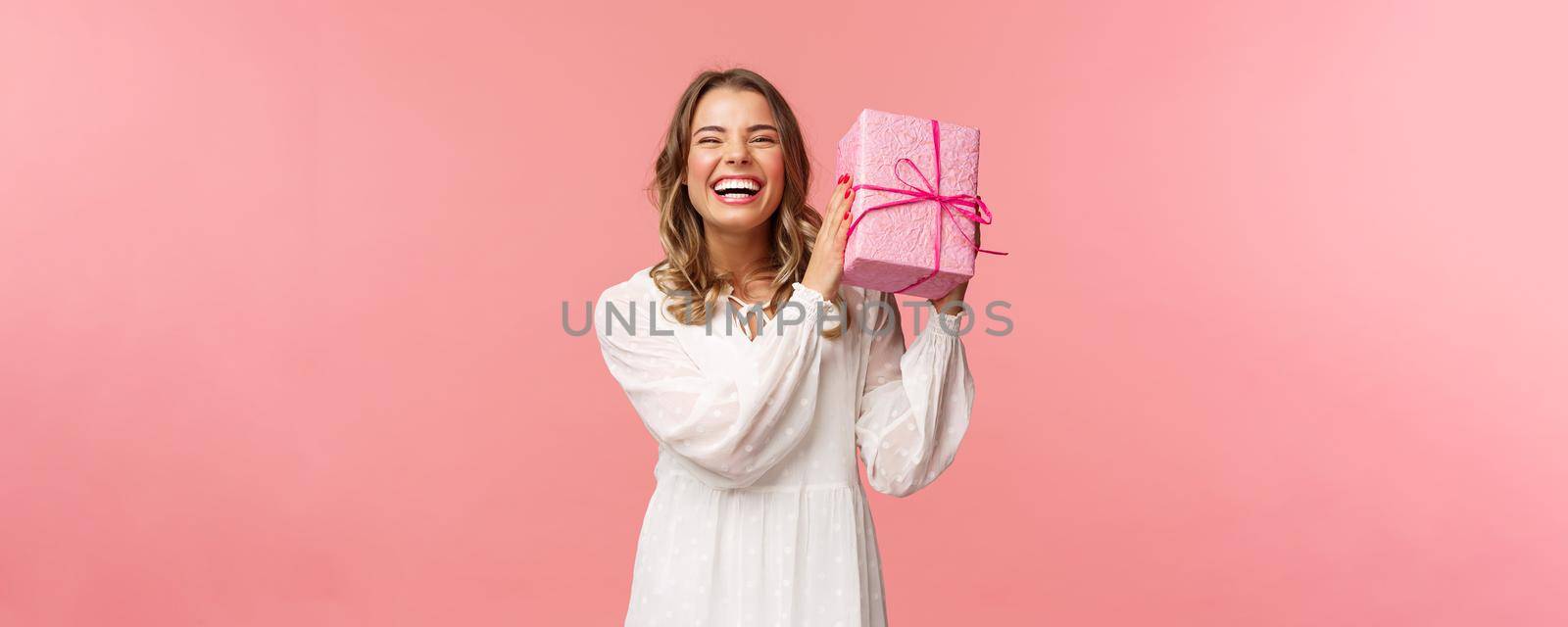 Holidays, celebration and women concept. Portrait of happy charismatic blond girl shaking gift box wondering whats inside as celebrating birthday, receive b-day presents, pink background.