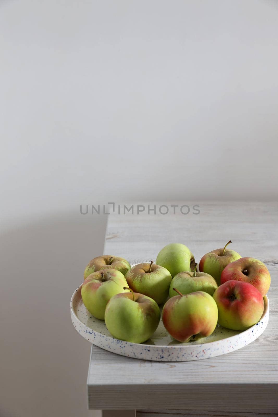 The green and red apples in a white faience bowl on the edge of a beige kitchen table. Scandinavian style. Minimalism. Place for text. by elenarostunova