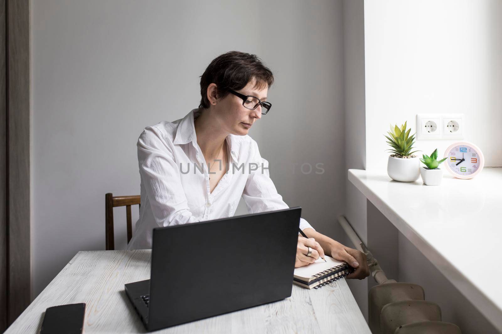 Middle-aged woman with short-haired brunette in glasses works at a laptop near window. She writes thoughtfully in a notebook