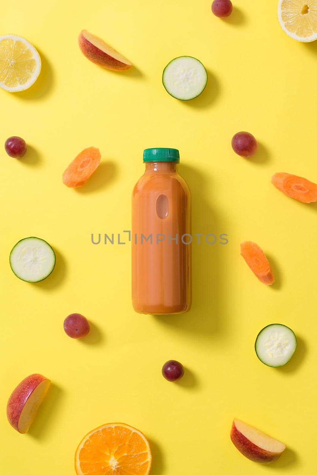 bottle of orange fruit and vegetable juice in the center of the image surrounded by pieces of fruits and vegetables of various colors on a yellow background