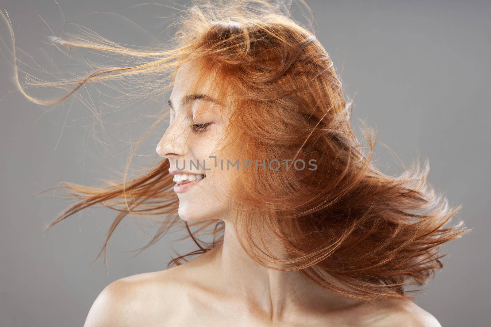 Beautiful dark burnt orange windy hair girl smiling. Studio portrait with happy face expression against gray background... by kokimk