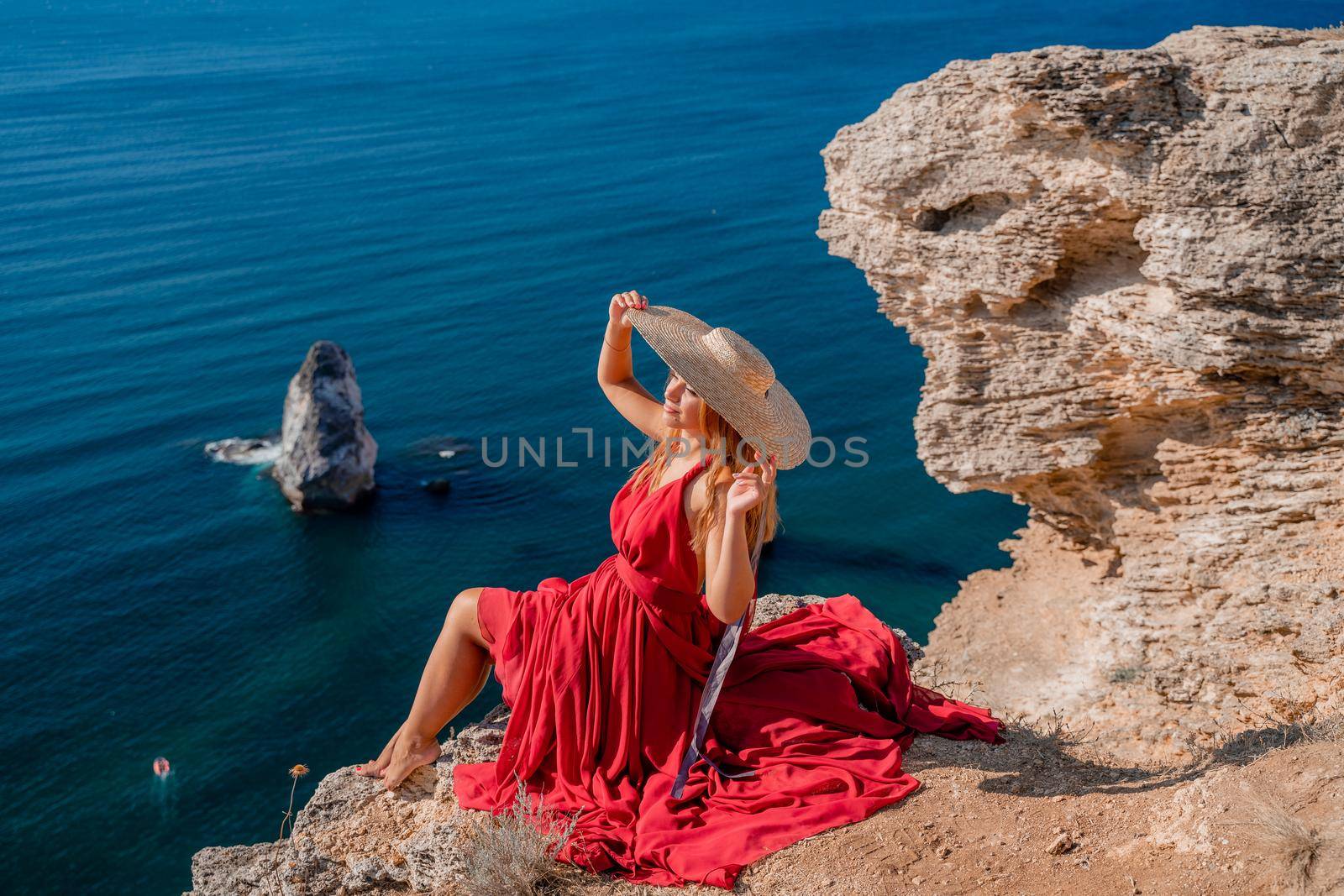 A girl with flowing hair in a long red dress sits on a rock above the sea. The stone can be seen in the sea. by Matiunina