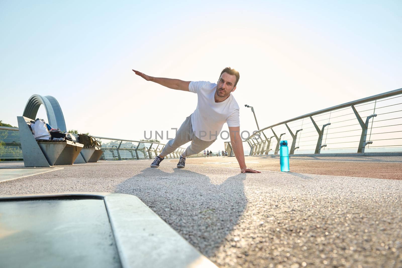 Active muscular European middle aged man, athlete, sportsman doing push-ups from the floor, alternately raising one arm during an outdoor workout at dawn on the city bridge