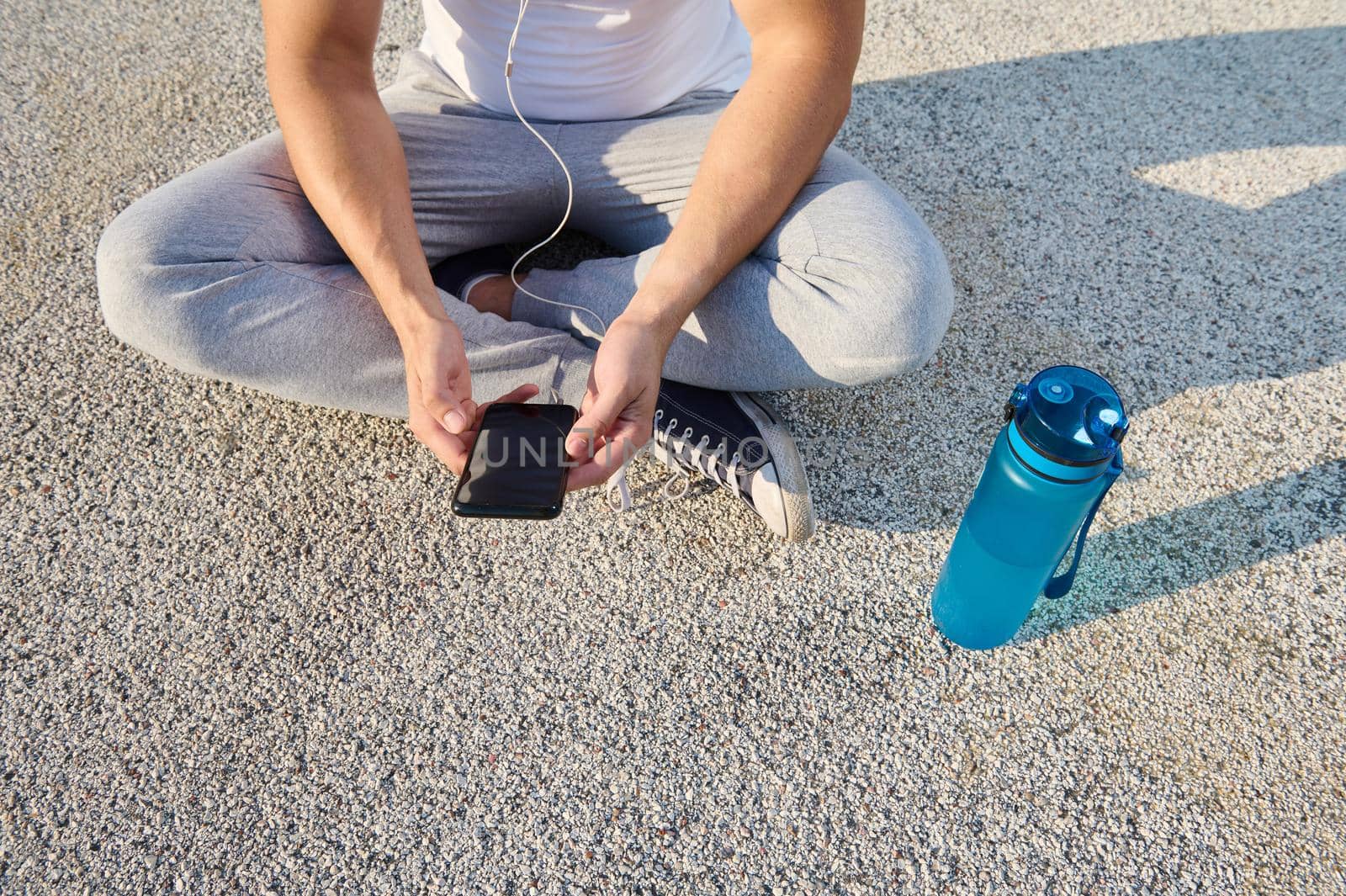 Overhead view of an athlete wearing earphones sitting on the city bridge and checking his phone while relaxing after morning cardio workout and using a fitness tracking mobile application