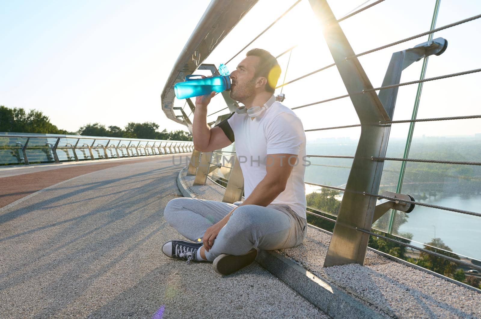 Side portrait of a muscular sportsman drinking water and relaxing after a heavy workout on a glass city bridge. The sunbeams of the early morning fall on him. by artgf