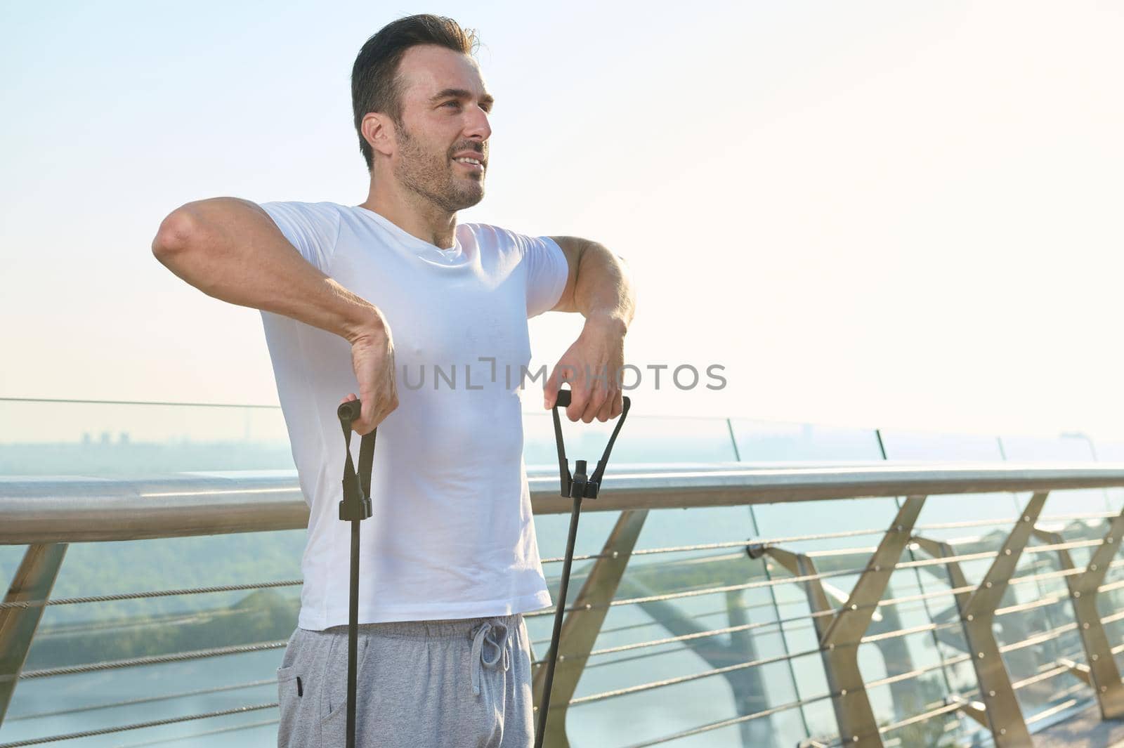 European young sportsman, athlete during outdoor workout with rubber resistance band. Middle-aged athlete doing exercises on shoulders and arms using an elastic expander on glass city bridge at dawn