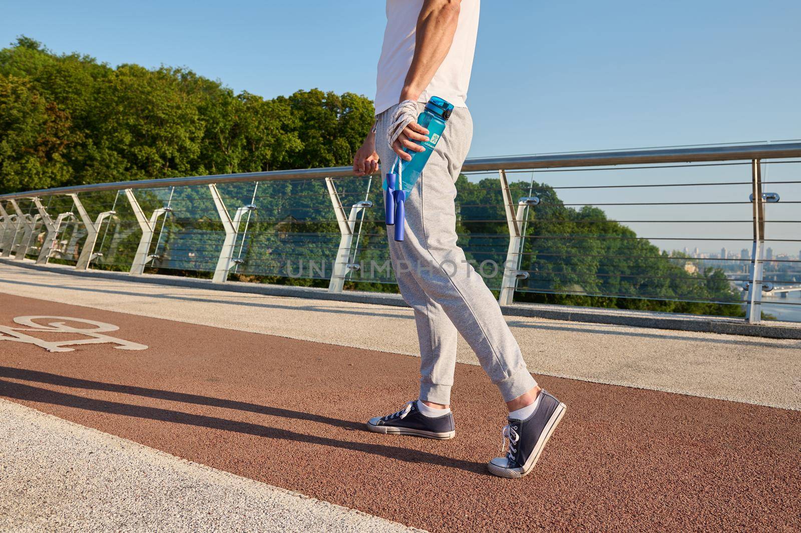 Cropped image sportsman holding a bottle with water and a skipping rope, standing on a city bridge treadmill during morning cardio workout. by artgf