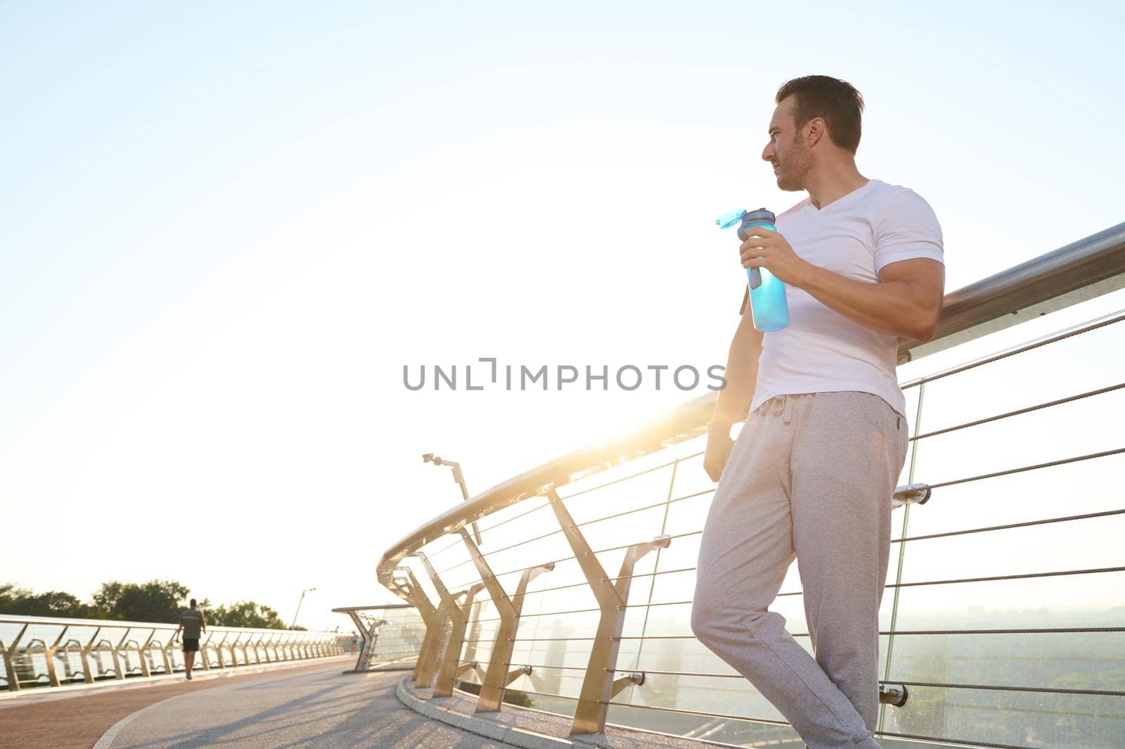 Full length portrait of a handsome sportsman athlete with a bottle of water, standing on the modern glass city bridge, relaxing after cardio workout outdoor. Sport, fitness, active lifestyle concept