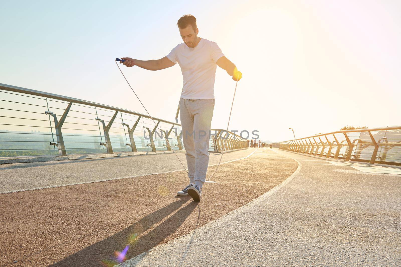 Full length portrait of a muscular build European middle aged man, fit athlete doing jumping exercises, cardio training with skipping rope on the urban city bridge early in the morning on a summer day by artgf