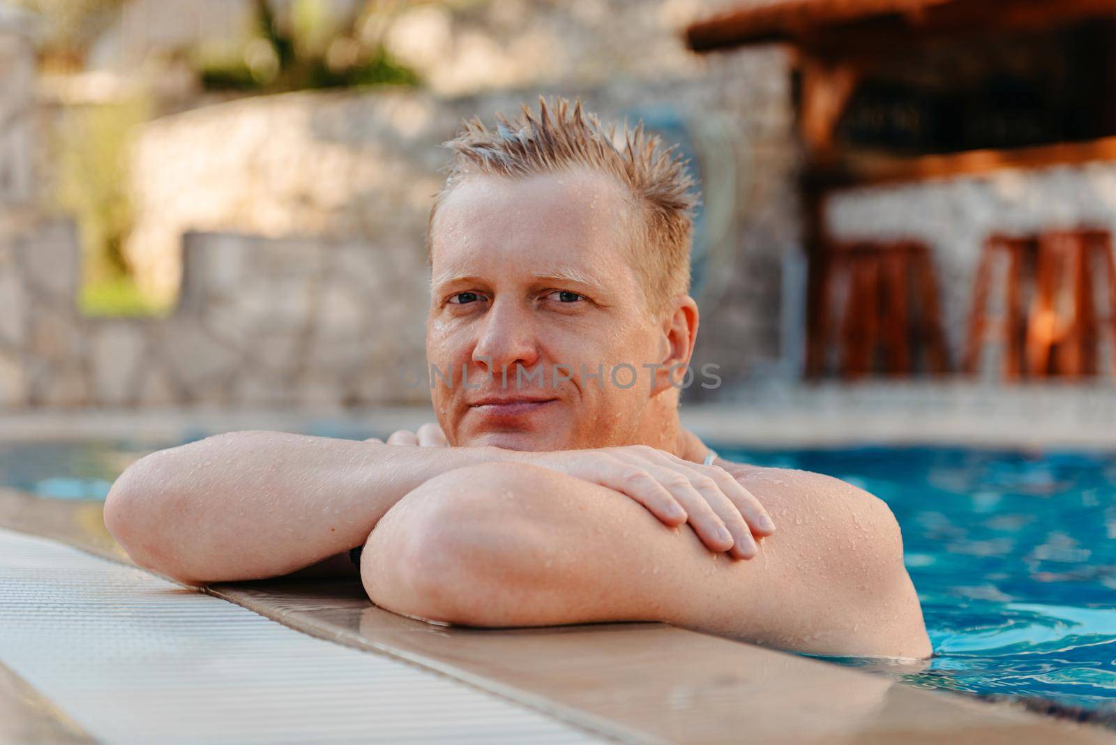 Photo of handsome smiling man in swimming pool in summer scenery. Attractive, cheerful, stunning, perfect, harsh, virile man in yellow shorts, having cocktail with small umbrella in hand and holding arm in pocket, isolated on blue background