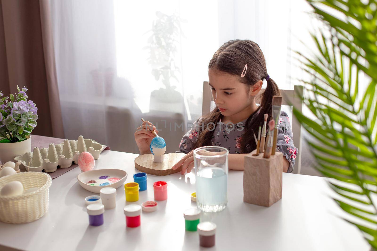 A little girl sits at a white table by the window, paints eggs with a brush and paint in jars. Copy space