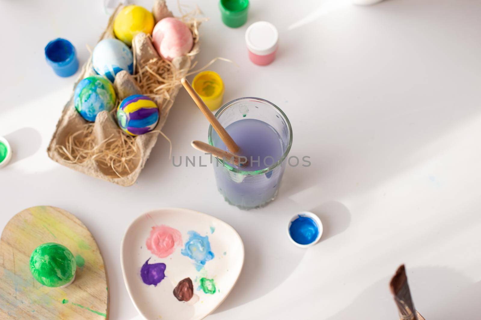 On a white table, in daylight, there are brushes in a glass of water, multi-colored eggs in a tray, jars of paint. Top view. Copy space