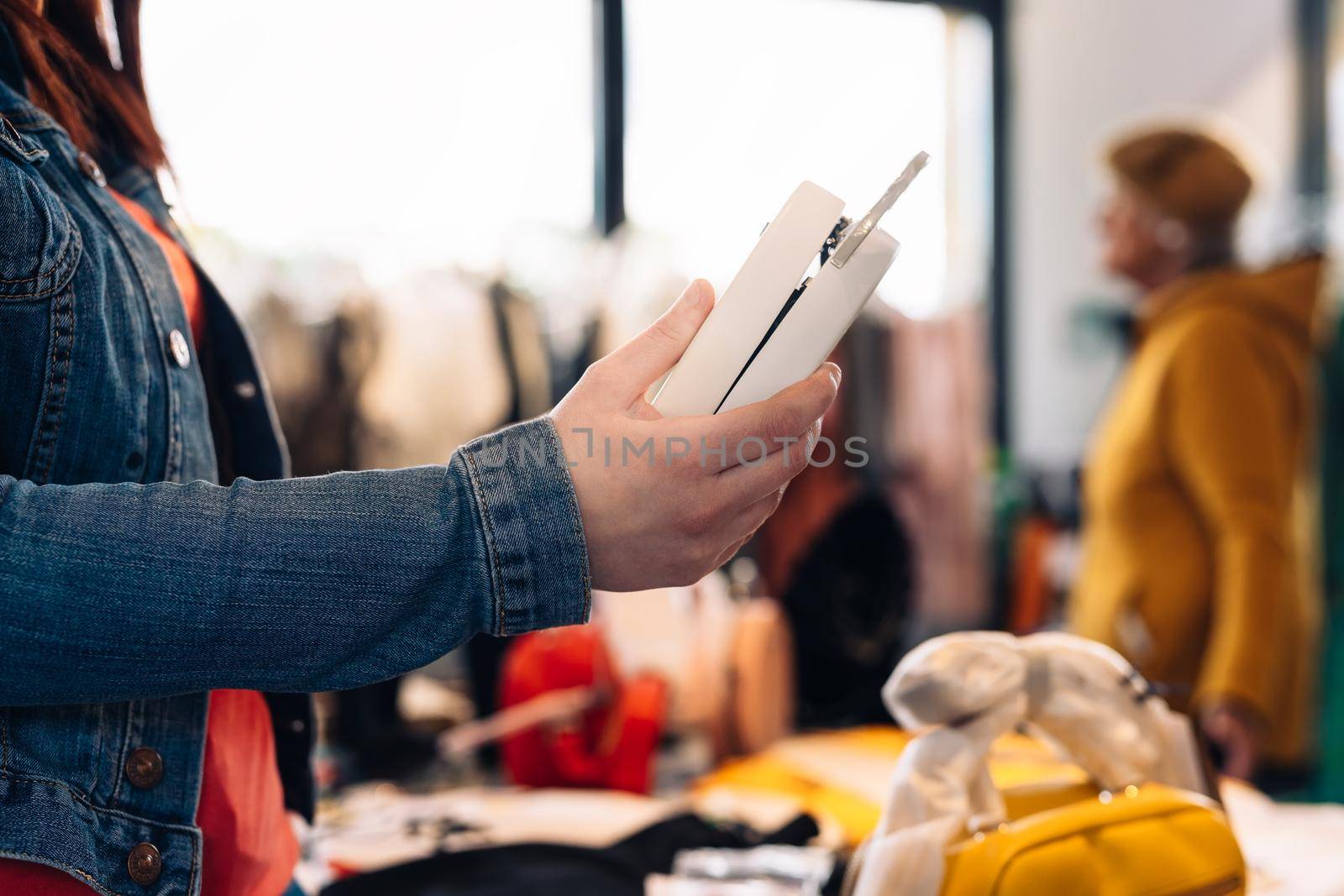 hands of a young woman, enjoying a shopping day, looking at a handbag to buy it, in a fashion shop. shopping concept. Natural light, sun rays, display with clothes, clothes rack, customers in background walking, clothes, Horizontal. customer with blue denim jacket, red t-shirt and jeans.