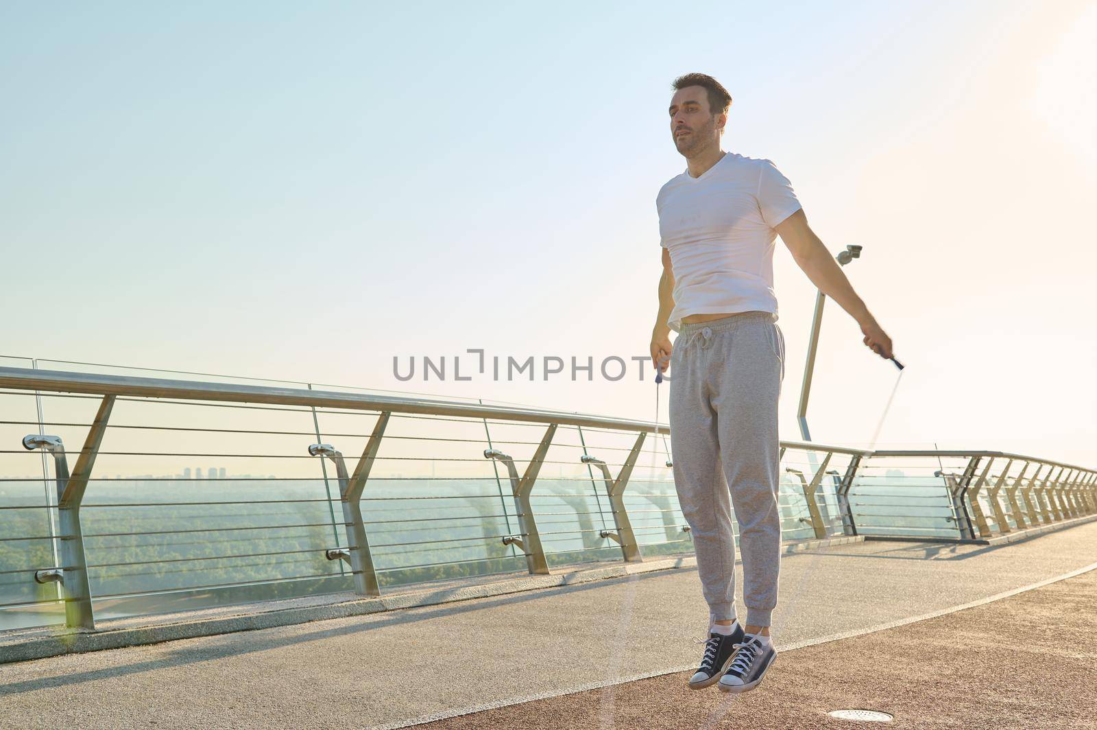 Full length portrait of a muscular build European middle aged man, fit athlete doing jumping exercises, cardio training with skipping rope on the urban city bridge early in the morning on a summer day