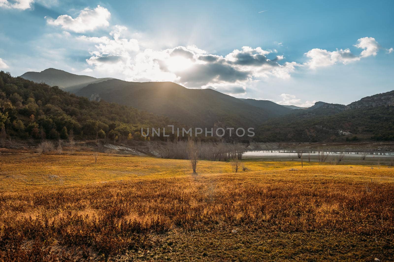 Dawn in the valley in autumn season with orange field and mountains around. Sun shines through the clouds on the blue sky.