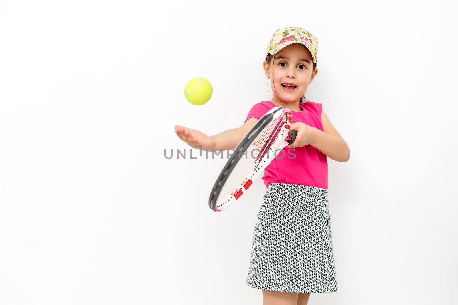 Studio shot of a little smiling girl dressed in pink T-shirt and white skirt - sportswear for tennis on a white background. She stands with a tennis racket and throws a tennis ball