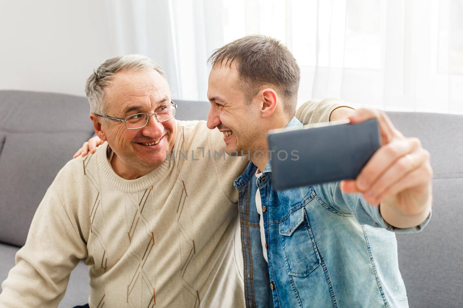 Happy moment. Cheerful young man taking a selfie with his upbeat elderly father waving at the camera and smiling pleasantly