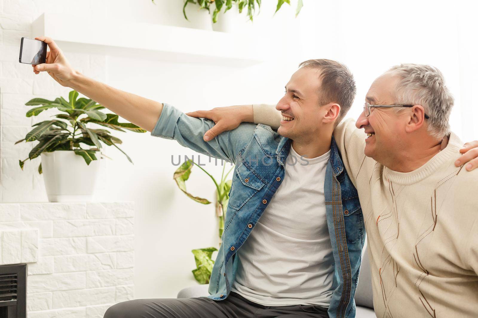 Happy moment. Cheerful young man taking a selfie with his upbeat elderly father waving at the camera and smiling pleasantly by Andelov13