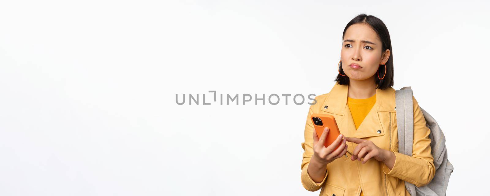 Image of happy girl traveller, tourist with backpack looking at smartphone, using mobile phone route app, standing over white background. Copy space