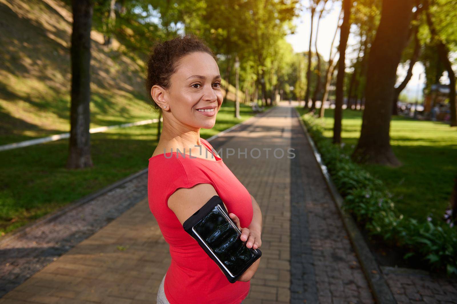 Portrait of a confident 40 years old African American woman in sportswear standing in forest city park with her arms crossed and smiling with a beautiful toothy smile during her morning run or workout by artgf