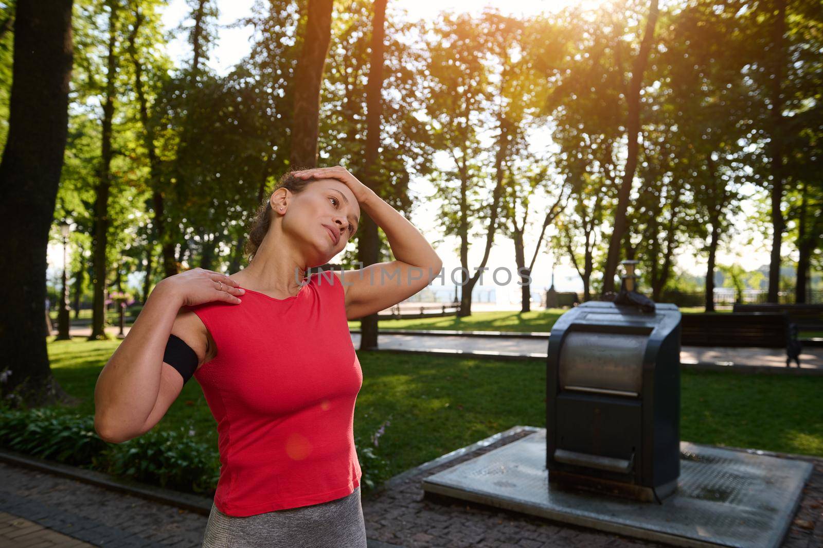 Waist length portrait of a charming African American young athlete, sports women doing stretching exercises while working out outdoor on a beautiful warm sunny summer day in the forest park by artgf