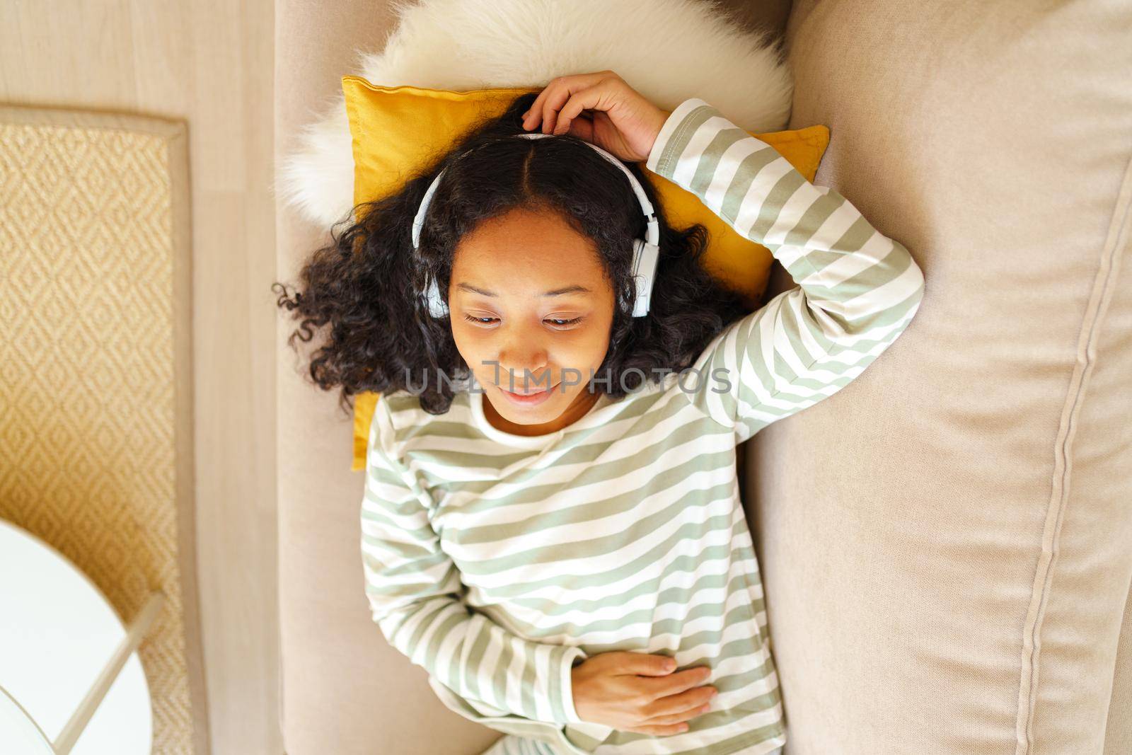 Calm African-American female listening to music while lying down on couch in living room, overhead by NataBene
