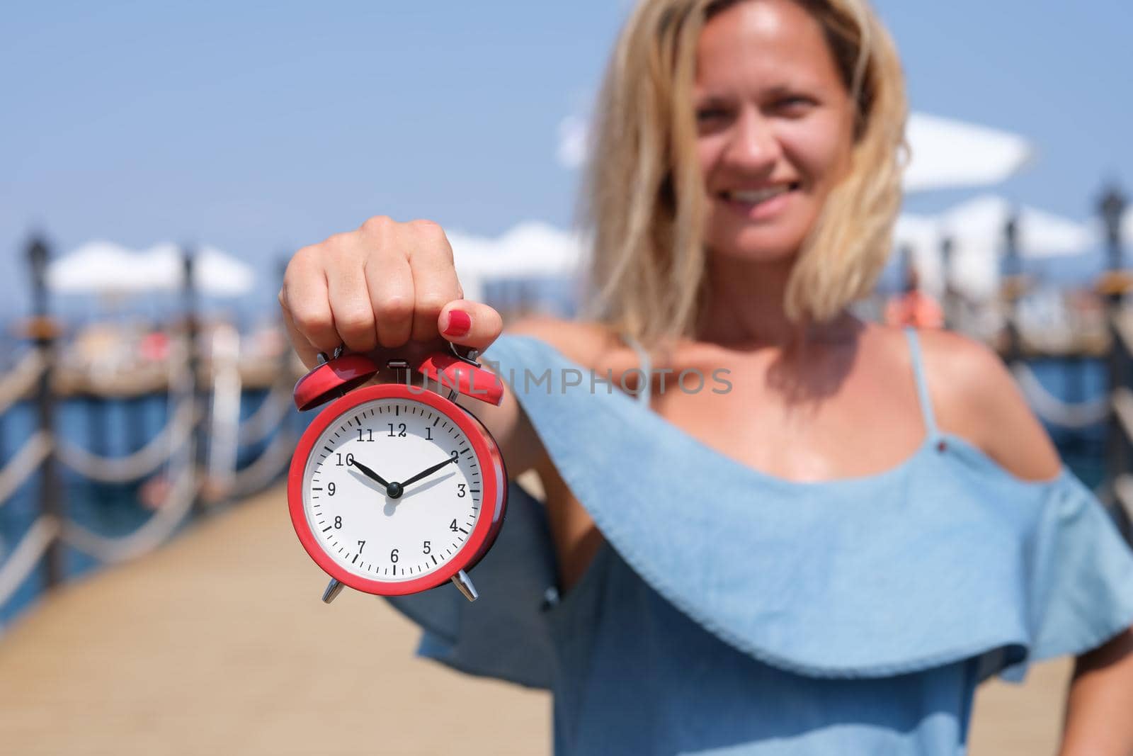 Smiling female tourist holding an alarm clock on sea pier. Travel and tourism concept