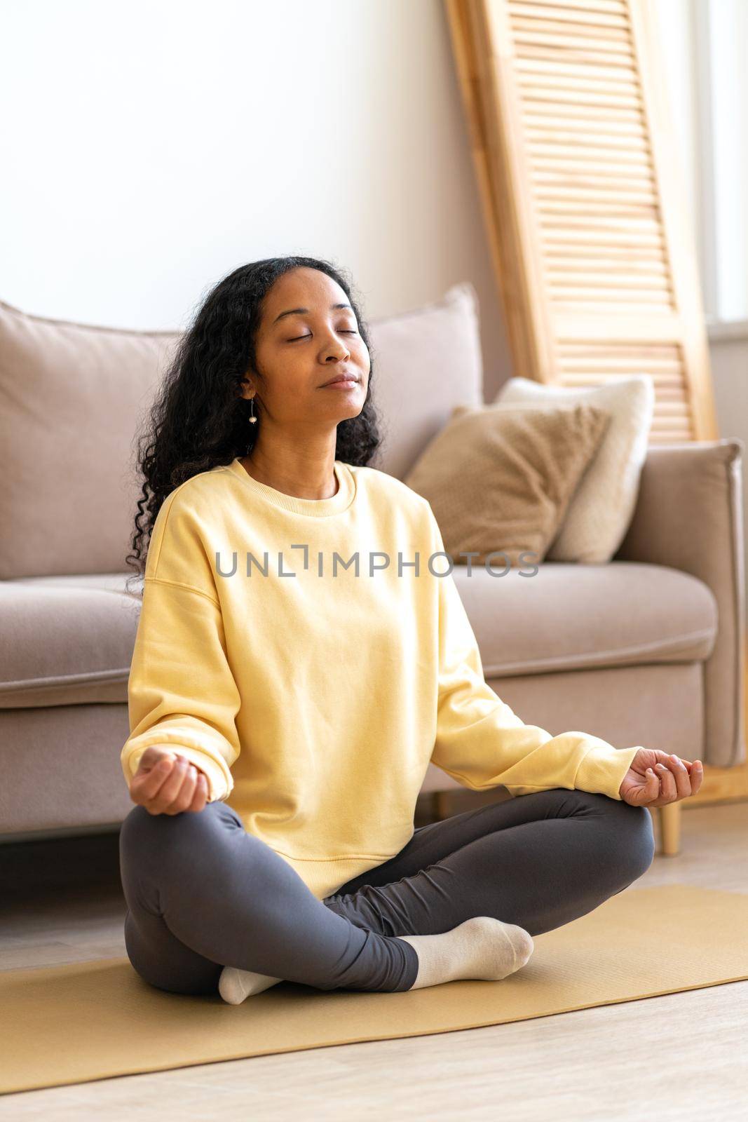 Young African-American female sitting in lotus yoga pose on mat in while meditating, vertical by NataBene