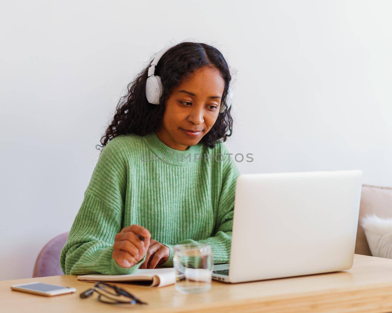 Young African-American female working on laptop in headphones while taking notes in notebook by NataBene
