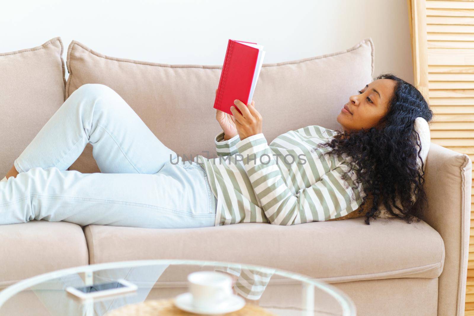 African-American lying down on sofa and reading book in red cover. Slow paced lifestyle by NataBene