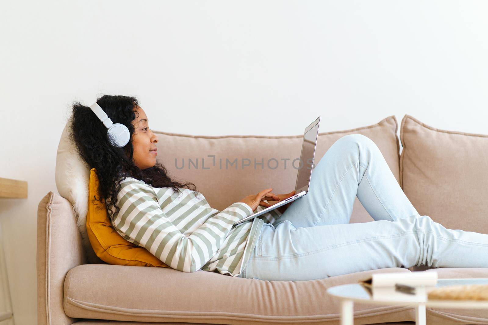 Young and smiling African-American female in headphones listening to music and chatting on laptop while lying on couch in lounge. Concept of spending leisure time online at home