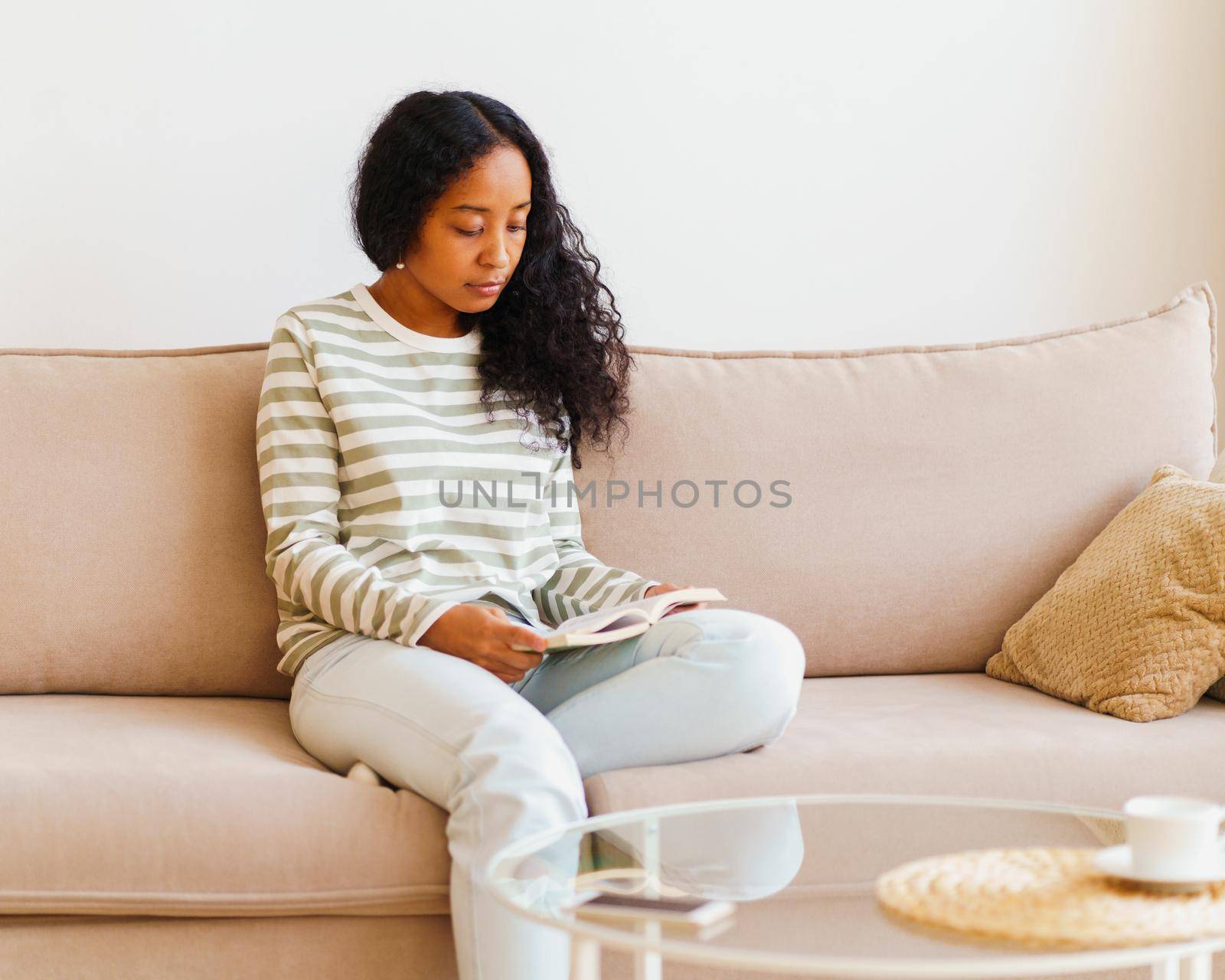 Beautiful African-American female sitting on couch and reading book. Spending time at home alone by NataBene