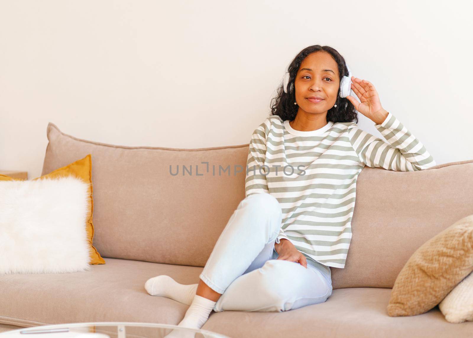 Smiling young African-American female in comfy clothes listening to music in wireless headphones while sitting on sofa in living room. Chilling and enjoying moment. Spending leisure time. Copy space