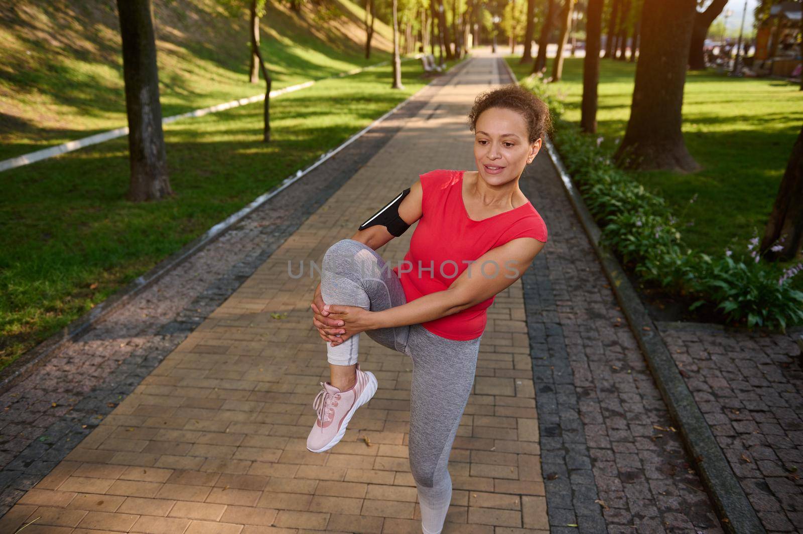 Determined cheerful African American fit woman in gray leggins and red tight t-shirt exercising outdoor in the city park. Fitness, body weight training outdoor on a warm summer day by artgf