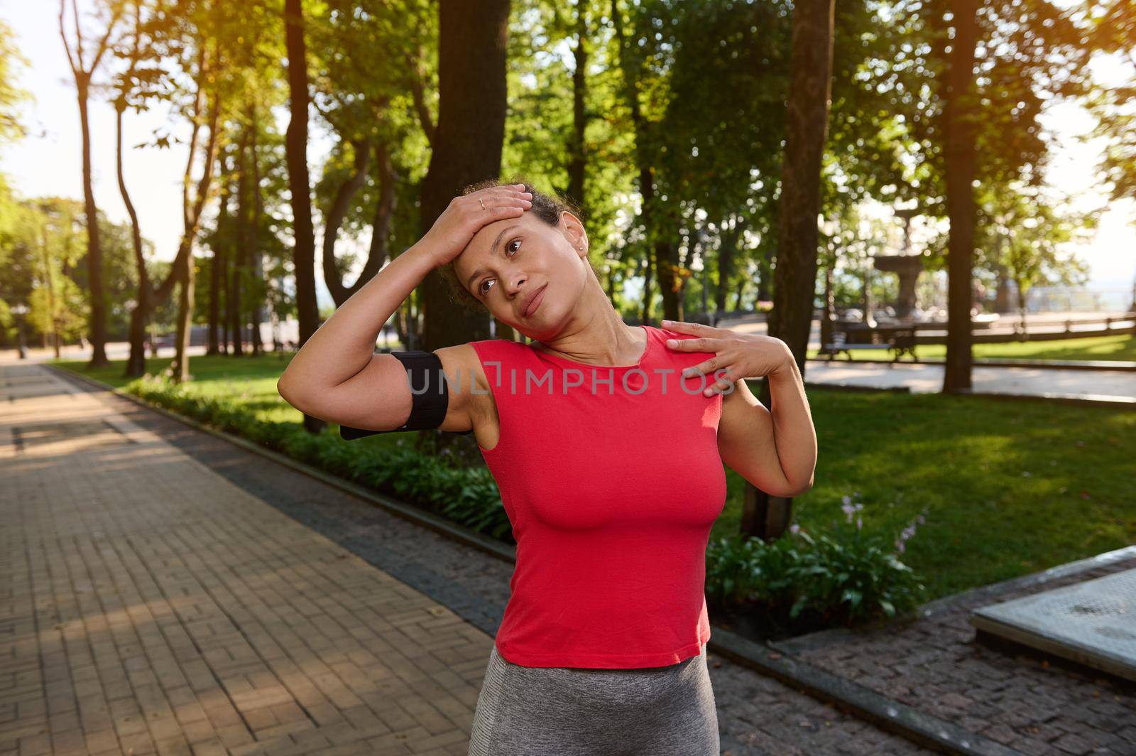 Hispanic sporty woman in tight red t-shirt with smartphone holder standing on the city park doing stretching exercise, enjoying morning workout or warming up her body before morning jog by artgf