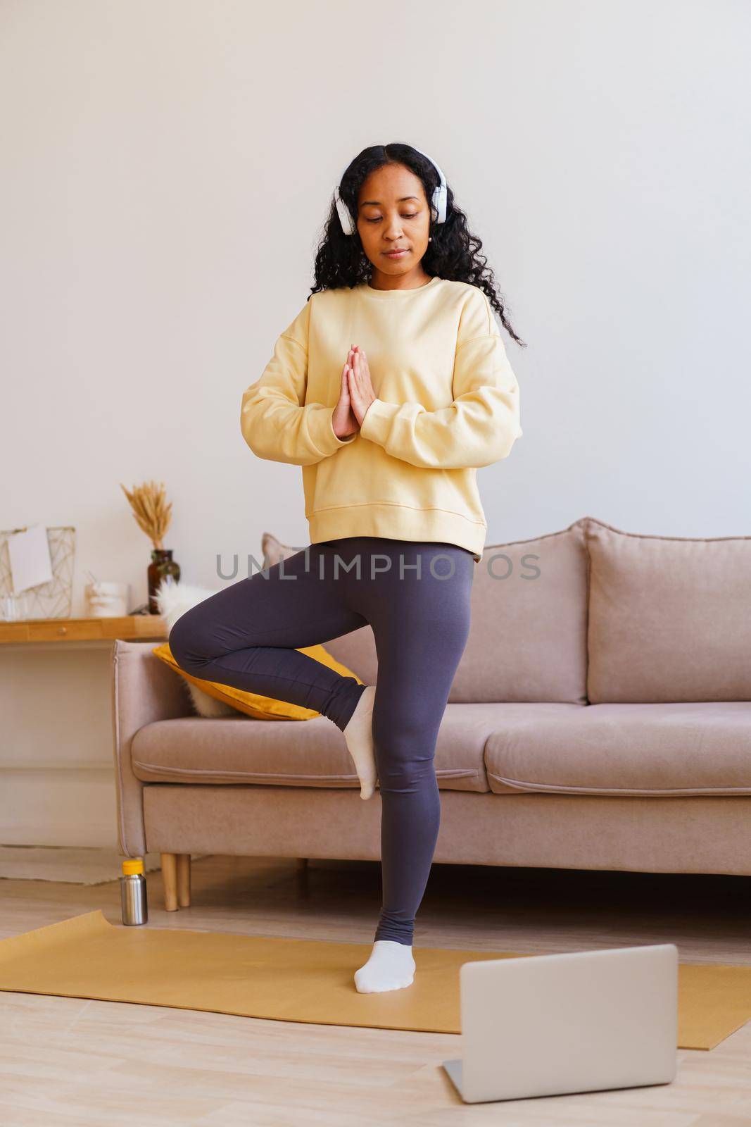Young African-American female in sportswear and headphones standing in yoga tree pose at home while following fitness tutorial on laptop. Vertical. Maintaining physical and mental wellbeing