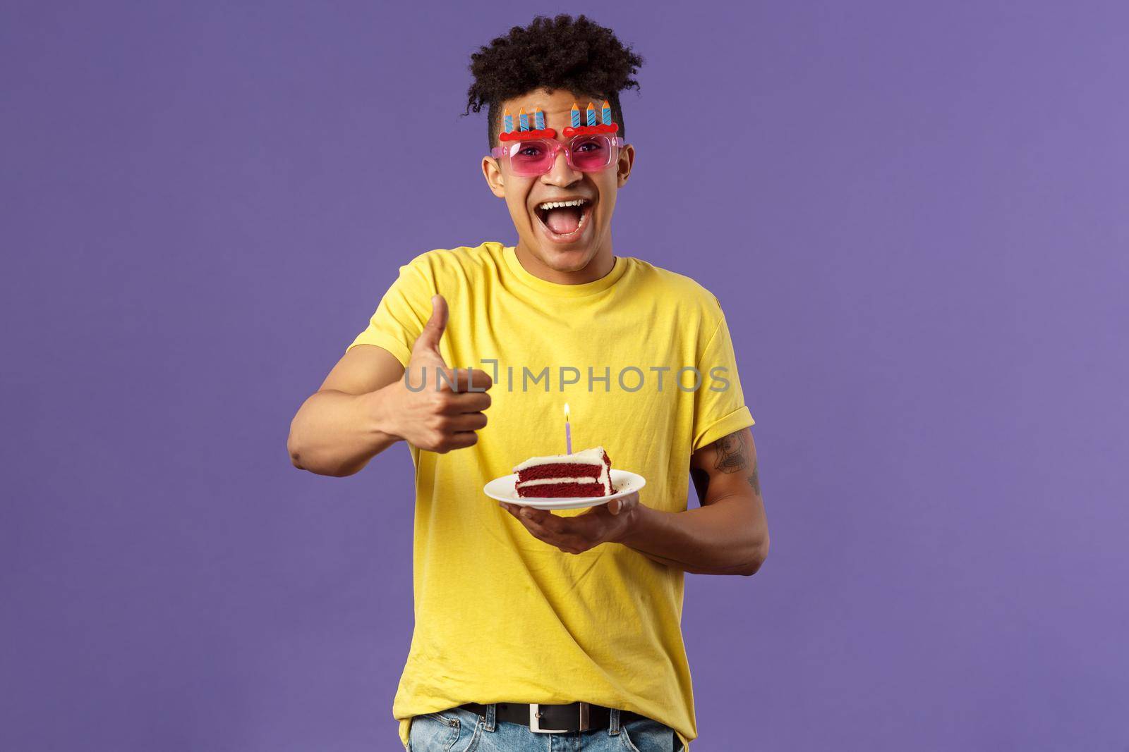 Celebration, party and holidays concept. Portrait of upbeat, cheerful young happy man celebrating birthday, wear funny sunglasses and hold b-day cake with lit candle, show thumb-up, recommend by Benzoix
