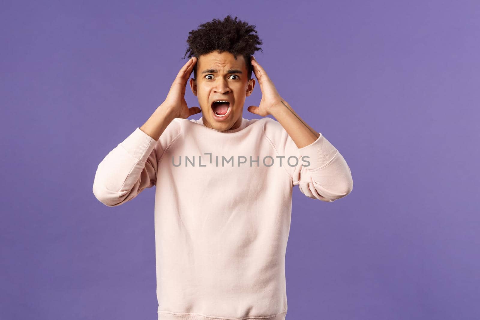 Portrait of scared and anxious young hispanic handsome man in panic, shaking hands nead head, screaming and gasping afraid, being in trouble, standing alarmed over purple background.