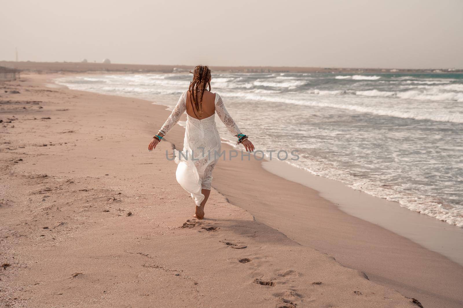 Model in boho style in a white long dress and silver jewelry on the beach. Her hair is braided, and there are many bracelets on her arms