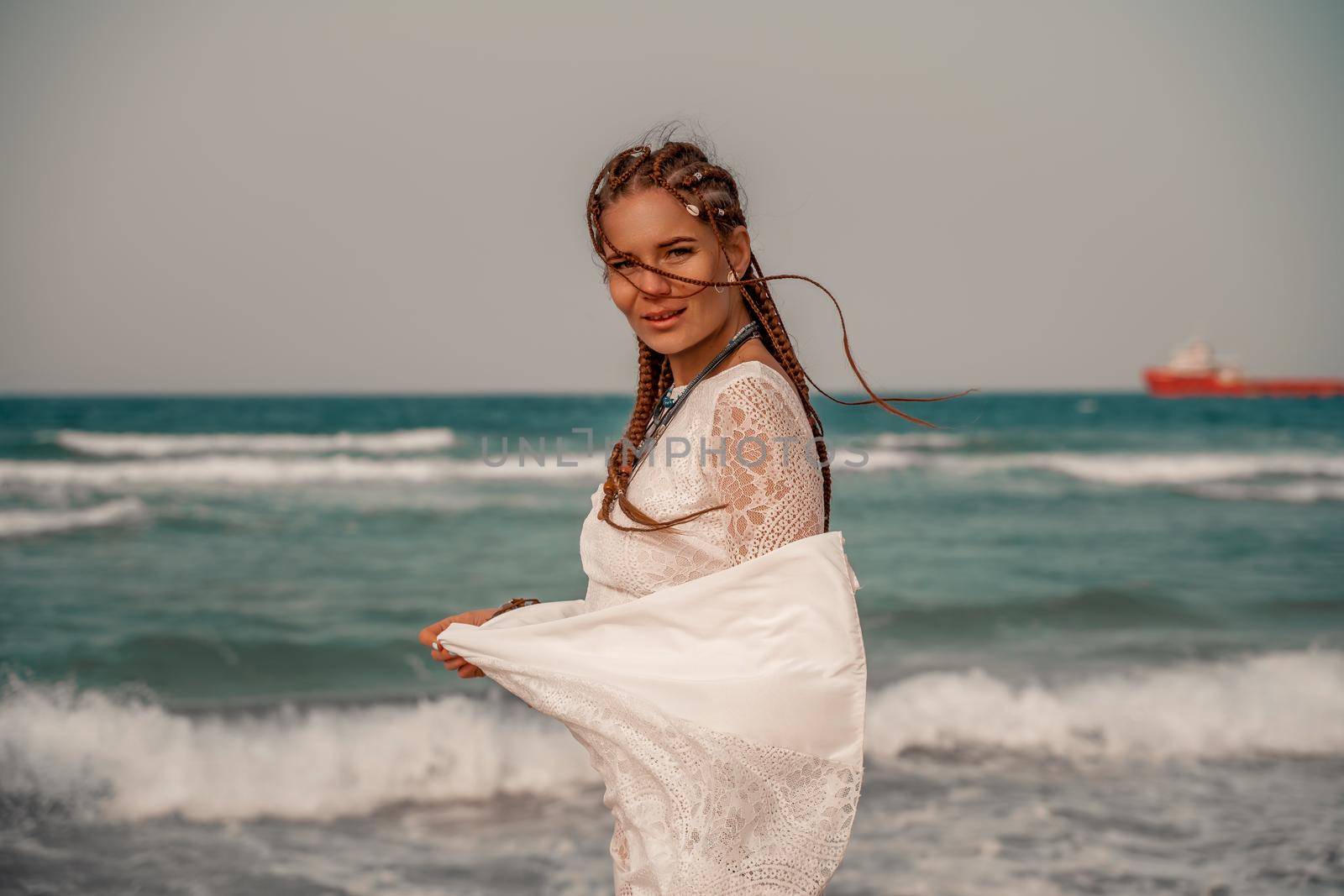 Model in boho style in a white long dress and silver jewelry on the beach. Her hair is braided, and there are many bracelets on her arms