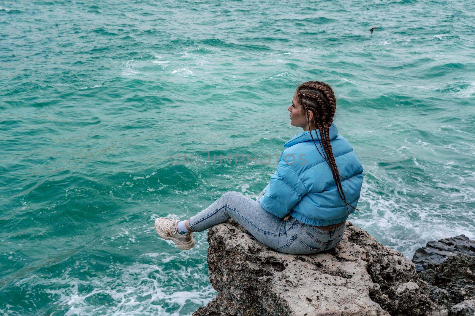 A woman in a blue jacket sits on a rock above a cliff above the sea, looking at the stormy ocean. Girl traveler rests, thinks, dreams, enjoys nature. Peace and calm landscape, windy weather