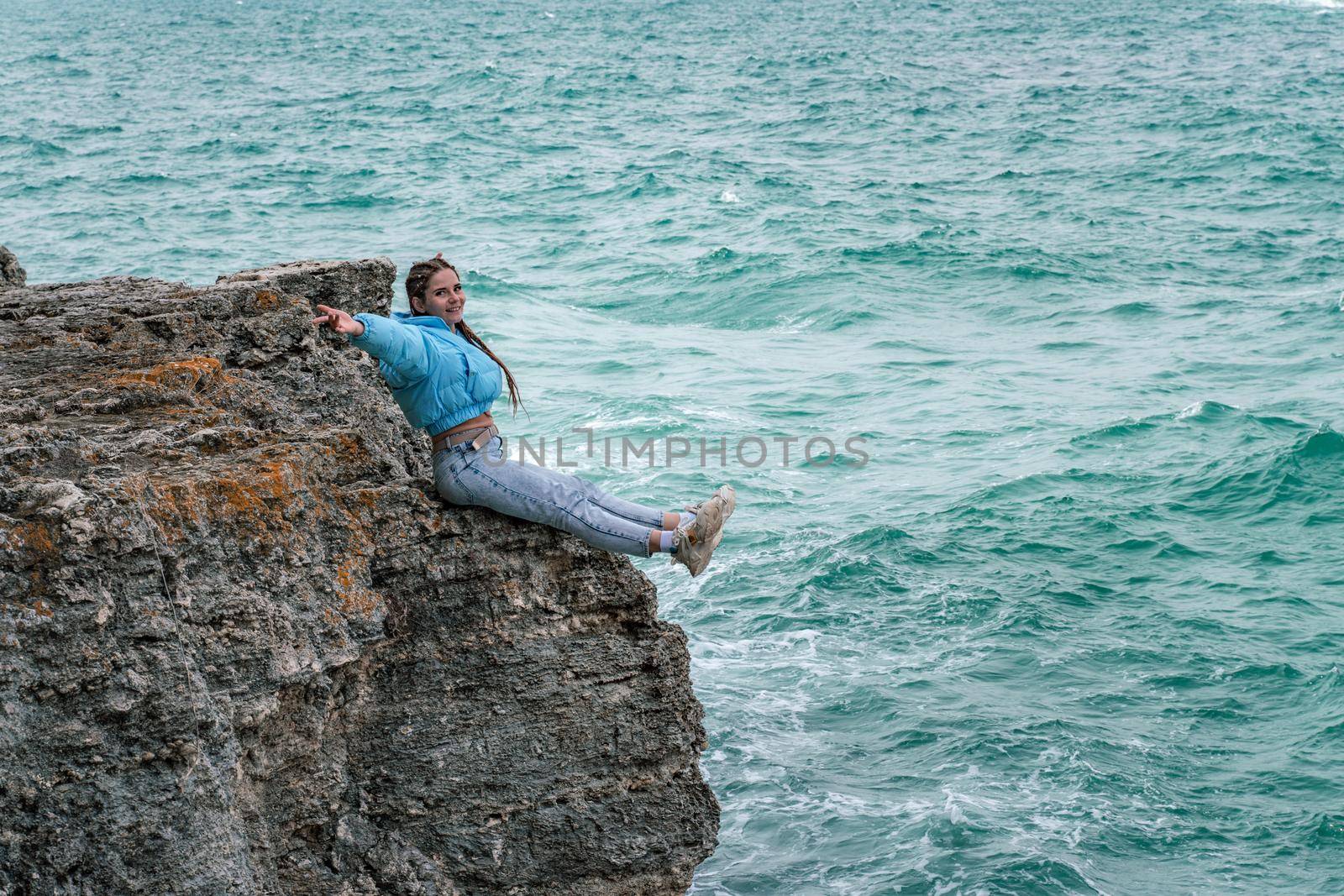 A woman in a blue jacket sits on a rock above a cliff above the sea, looking at the stormy ocean. Girl traveler rests, thinks, dreams, enjoys nature. Peace and calm landscape, windy weather