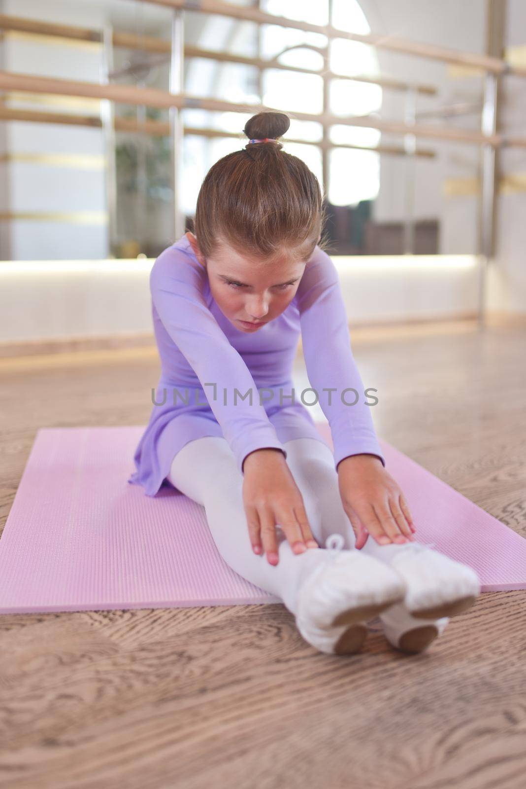 Beautiful little ballerina in a lilac leotard and white tights doing stretching exercises on a pink karemat in dance studio. by Nickstock