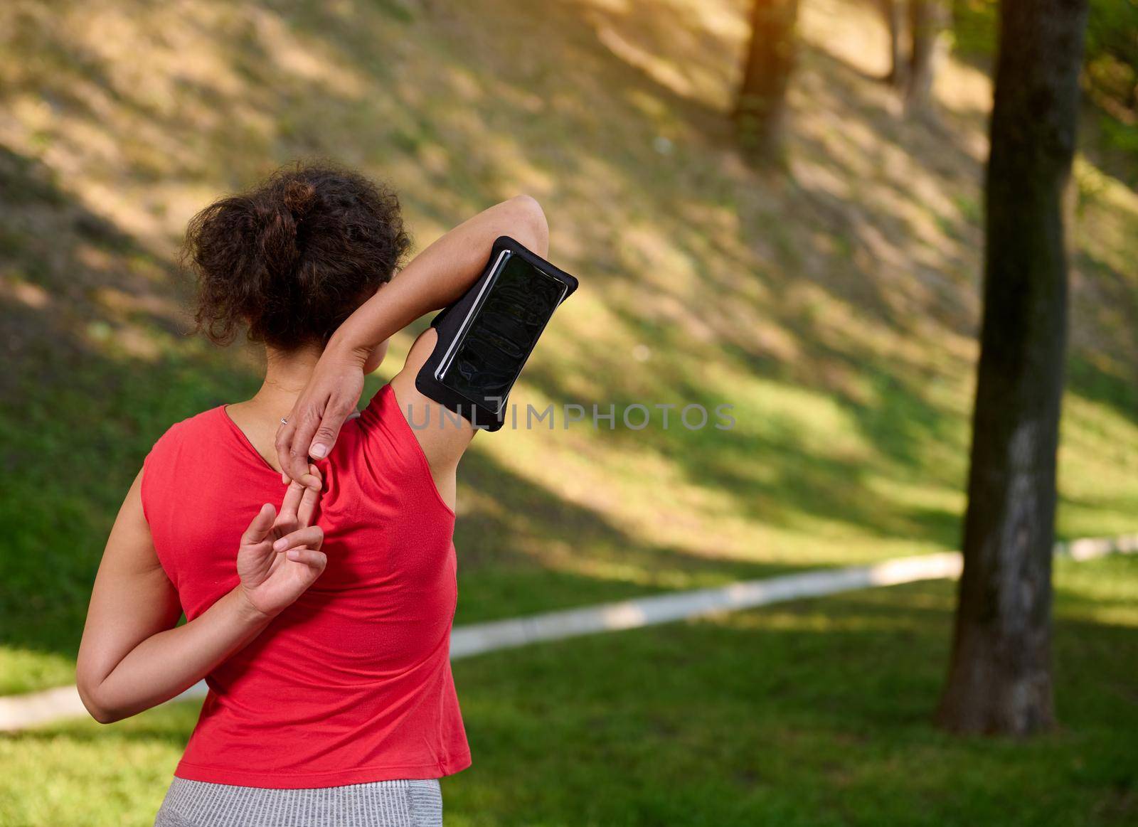 Rear view of an African American woman, athlete in sportswear, wearing a smartphone holder, exercising outdoor, warming up and stretching out her body during morning workout by artgf