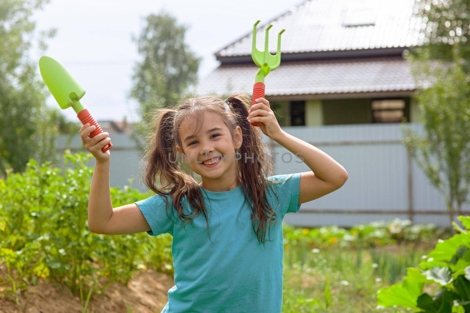 Cute little girl in green t-shirt holding small gardening tools , dancing in the garden on a sunny day. Copy space