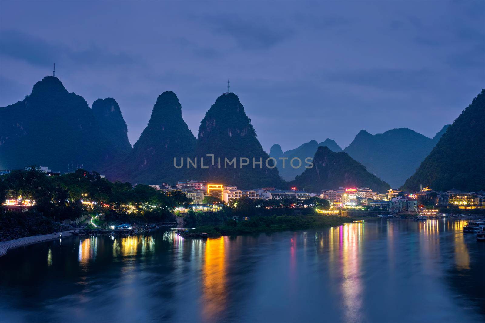 View of Yangshuo town illuminated in the evening with dramatic karst mountain landscape in background over Li river. Yangshuo, China