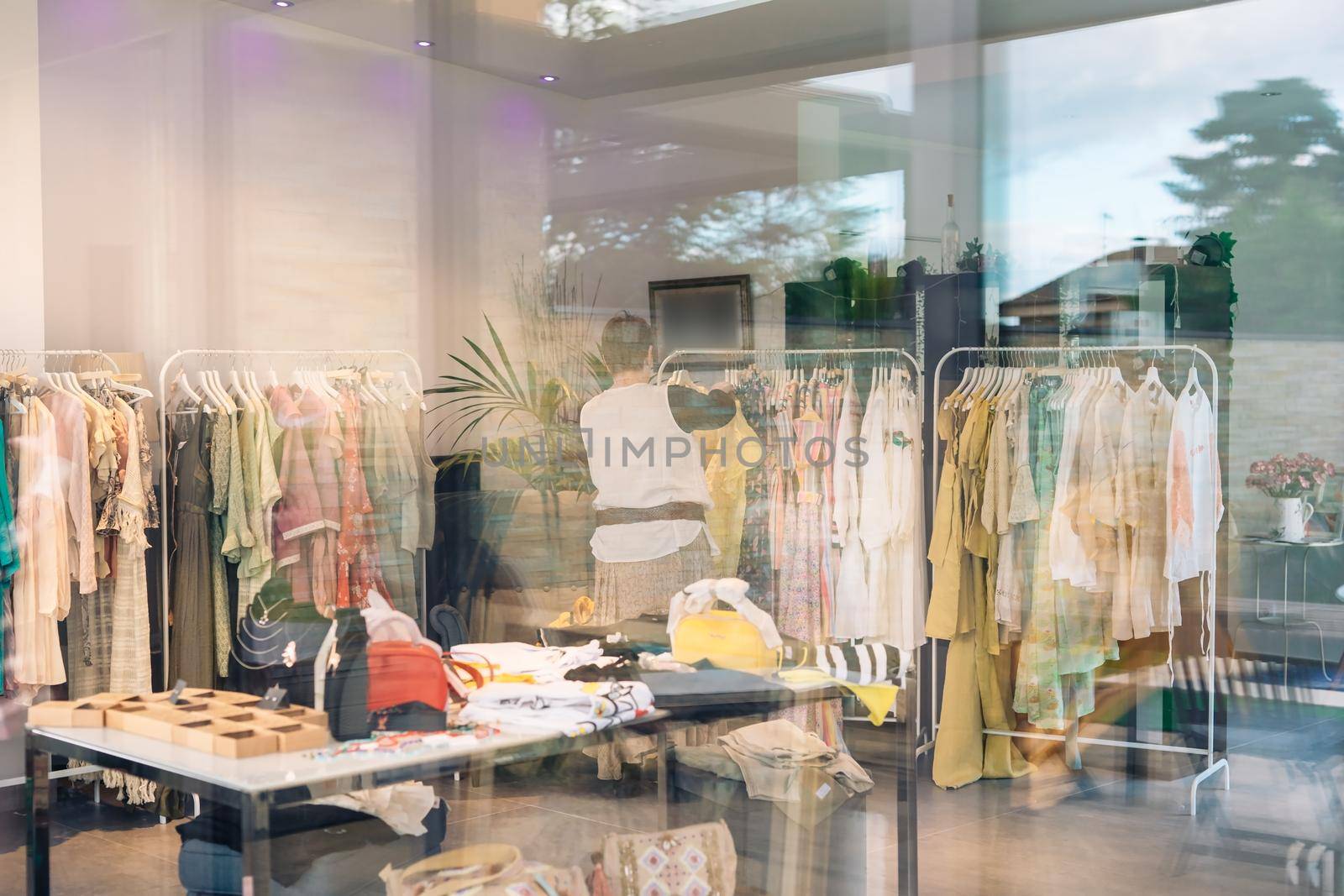 view from the outside of an elderly woman inside a clothes shop. shop window, shopping concept. woman selling clothes in her small shop. natural light from the window, sun rays, mirror, display with clothes, coat rack, window, clothes, Horizontal.