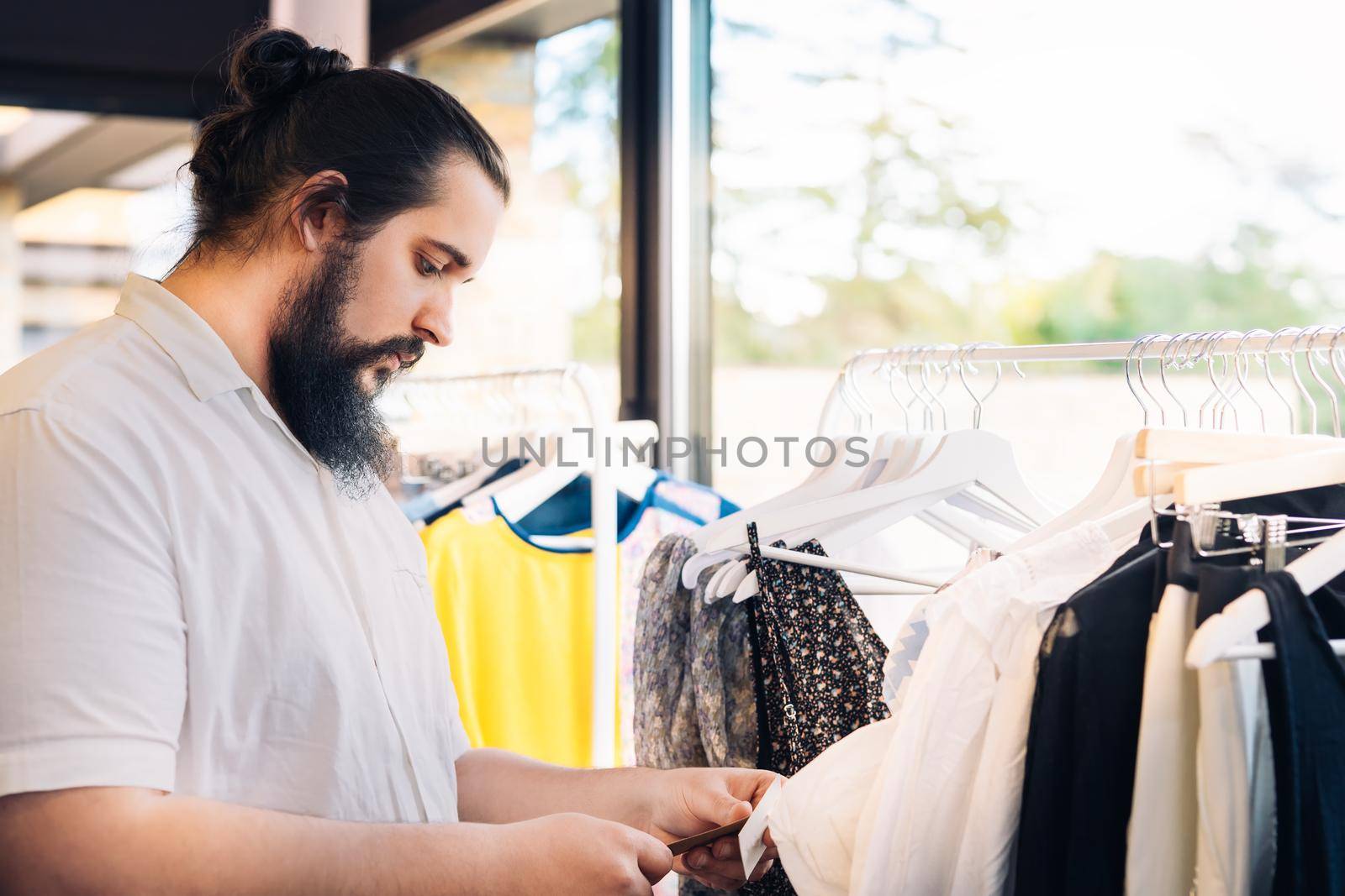 young man with beard, looking and buying clothes, in fashion shop. shopping concept. leisure concept. Natural light, sunbeams, clothes rack with coloured clothes, horizontal view, copy space. dress with white shirt.